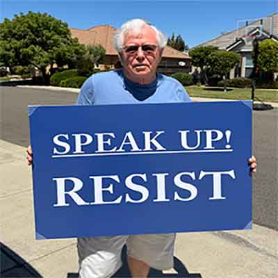 An older man alone holding protest sign that says Resist.